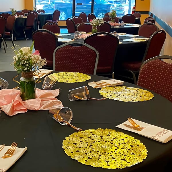 Restaurant dining room with black-draped tables, red chairs, and gold place settings with floral centerpieces.