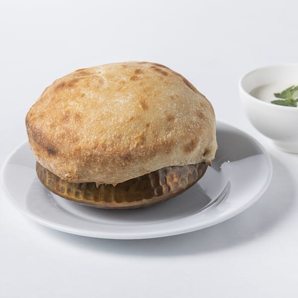 A golden-brown puffed bread, likely a Nepalese roti, on a white plate with a small bowl of sauce.