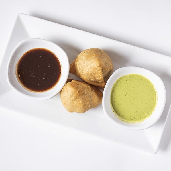 Two golden-brown samosas on a white plate, served with dark and light green dipping sauces.